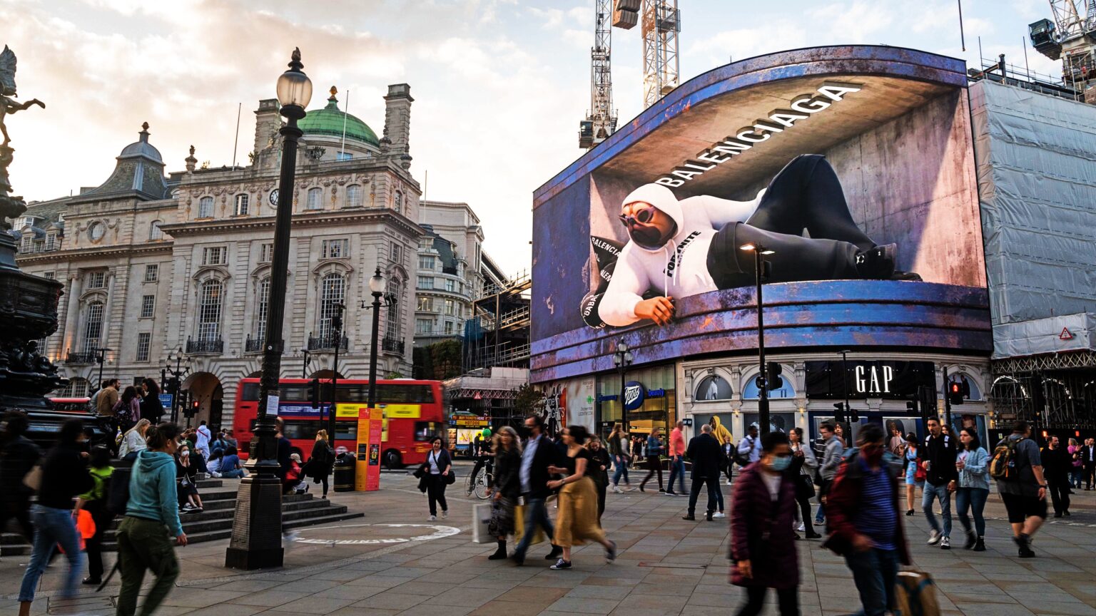 Large digital billboard in an urban square featuring a Balenciaga advertisement with a model lounging in streetwear.