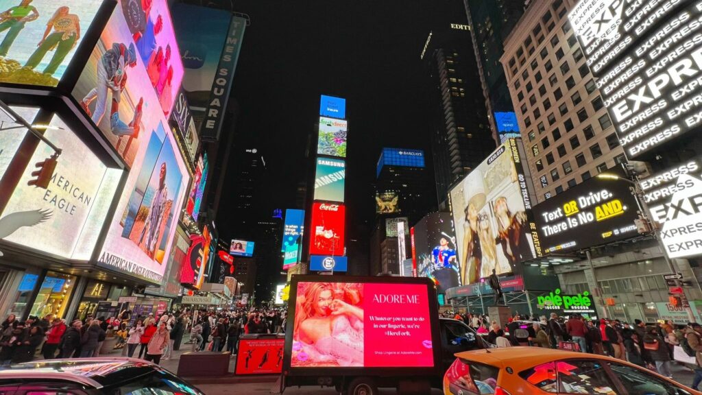 Times Square at night, filled with crowds and illuminated by large digital billboards from brands including Samsung, Coca-Cola, American Eagle, and Adore Me