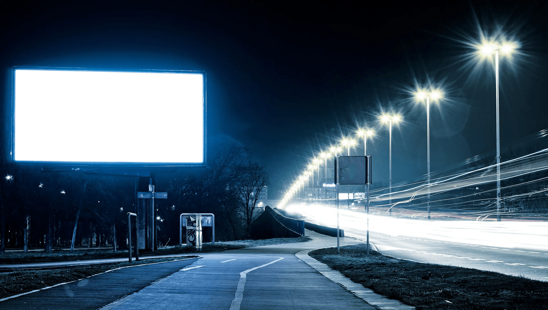 Illuminated blank billboard at night with streetlights and motion-blurred car lights on a city road.