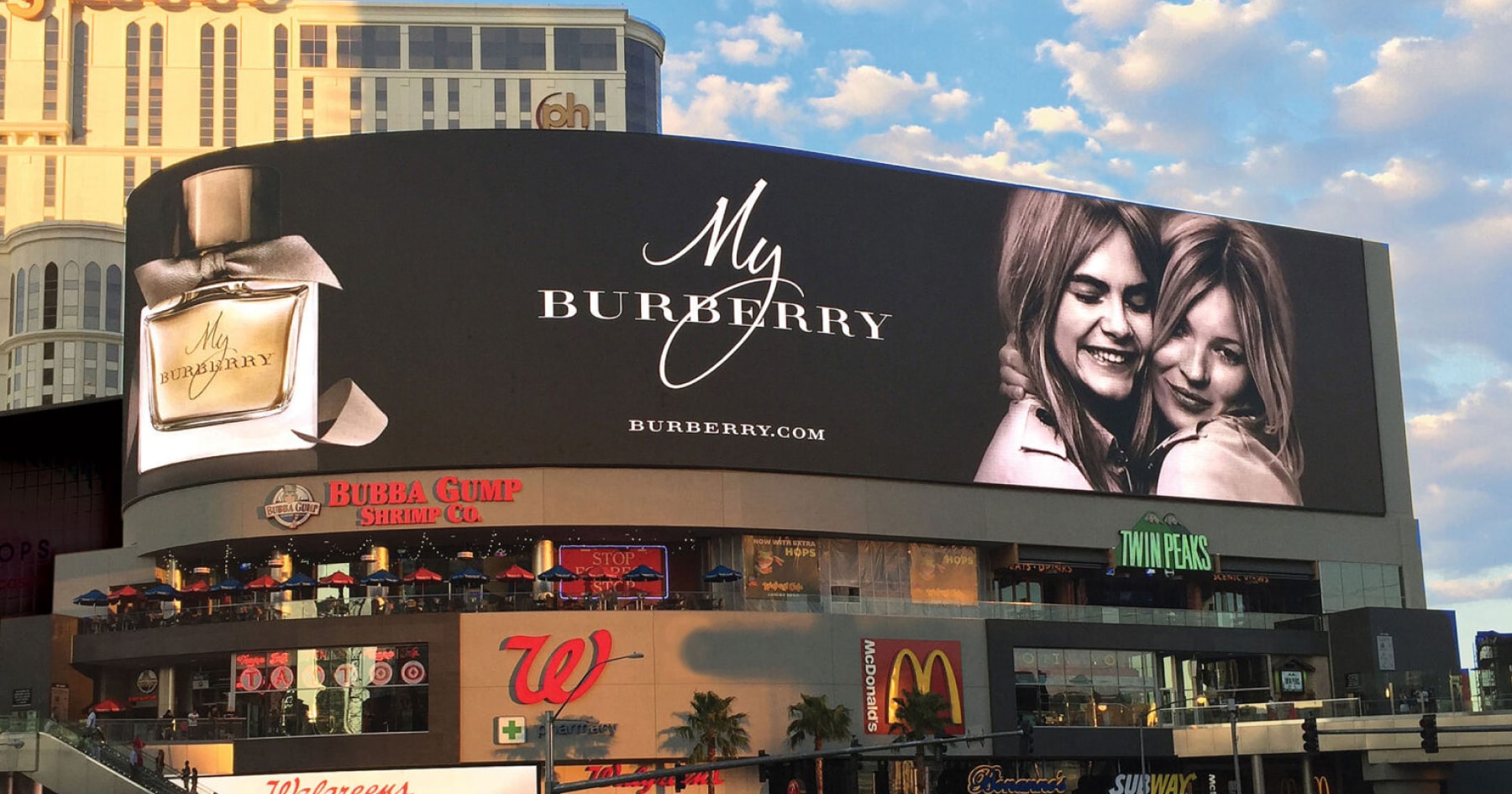 Outdoor advertisement for Burberry perfume on a digital screen, showing a perfume bottle and two models, located above retail stores and eateries.